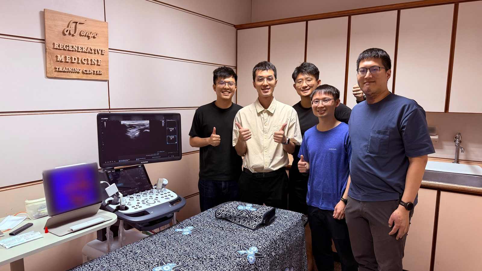 五人在超音波教室比讚合照,旁邊有超音波機與鋪著花布的治療床,牆上掛著訓練中心的木牌
